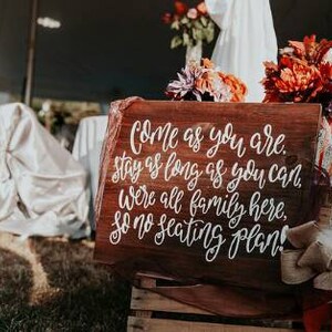 May include: A rustic wooden sign with white calligraphy that reads "Come as you are. Stay as long as you can. We're all family here, so no seating plan!"