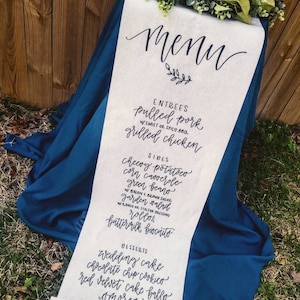 May include: A white scroll with a black handwritten menu for a wedding or event. The menu includes entrees, sides, and desserts. The entrees are pulled pork and grilled chicken. The sides are cheesy potatoes, corn casserole, green beans, garden salad, rolls, and buttermilk biscuits. The desserts are wedding cake, chocolate chip cookies, red velvet cake balls, and more.