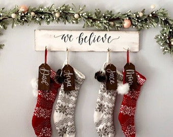 May include: A Christmas display featuring four knitted stockings, two red and two gray, each with a name tag. The stockings hang below a wooden sign that reads "we believe" in cursive, with a garland of greenery above.