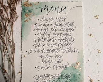 May include: A handwritten menu on textured paper with watercolor accents and gold leaf details. The menu lists dinner rolls, salad, grilled asparagus, twice baked potatoes, and other dishes. The word "menu" is at the top.