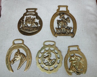 May include: Five brass horse brasses featuring equestrian motifs. One shows a group of riders, another a rider with hounds, two depict horse heads within horseshoe frames, and one is a circular medallion with a central prancing horse figure.