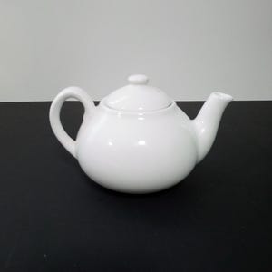 May include: A white ceramic teapot with a lid. The teapot has a round body and a spout on the side.