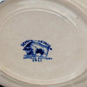 May include: A white ceramic plate with a blue rim and a blue buffalo logo. The logo includes the words "Semi-Vitreous" and "Buffalo Pottery 1911". The plate is likely vintage.