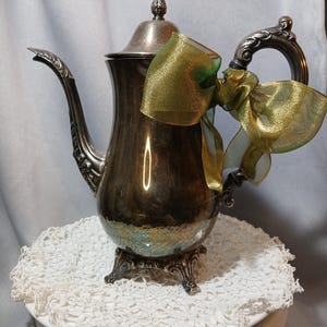 May include: A silver-toned metal teapot featuring an ornate handle, a curved spout, and a pointed lid. The teapot is decorated with a translucent, shimmering green and gold ribbon tied in a bow around the handle and the upper body of the vessel.