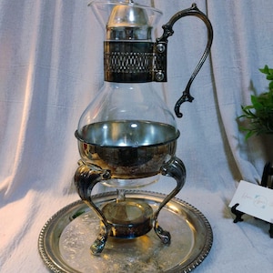 Peut inclure: Un ancien service à café ou à thé argenté avec une carafe en verre. Le service comprend une poignée décorative, un support chauffant et un plateau assorti. Le design présente des détails ornés et une esthétique classique.