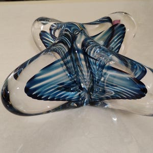 May include: A clear glass decorative object with a swirling blue design. The object has a unique, abstract shape, with rounded edges and a central knot-like feature. The blue design is intricate, with streaks and patterns throughout the glass.