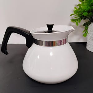 May include: A white coffee pot with a black handle and lid. The pot has a silver band around the middle and a spout for pouring. The pot is sitting on a black surface.