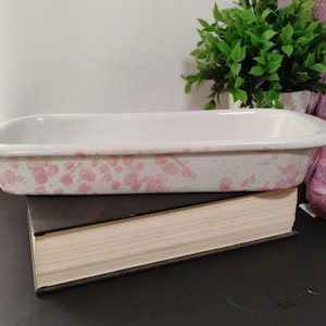May include: A rectangular white baking dish with pink splatter pattern. The dish is resting on a thick, dark-covered book. The dish has a slightly curved rim and is empty. A green plant is in the background.