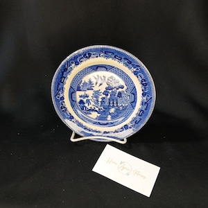 May include: A blue and white ceramic plate with a Willow pattern design. The plate is round and features a detailed landscape scene in the center, surrounded by a decorative border. The plate is displayed on a white metal stand.