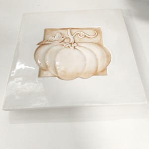 May include: A square, white ceramic decorative tile with a raised relief of a pumpkin. The pumpkin is tan with a stem and leaves, set against a rectangular tan background. The tile is likely for seasonal or fall decor.