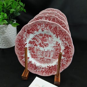 May include: A set of four ceramic plates featuring a red and white transferware pattern of a castle and deer. The plates have scalloped edges and a floral border. They are displayed on a wooden stand with a white card labeled Home Again Interiors.