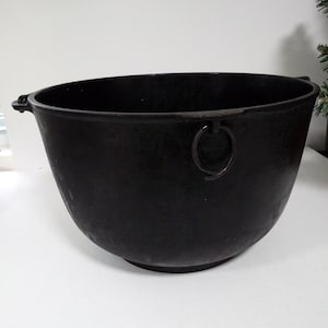 May include: A large, black cast iron cauldron with a rounded shape and two small handles on the sides. A metal ring is attached to the side of the cauldron. The cauldron is sitting on a white surface.