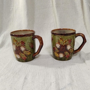 May include: Two ceramic mugs with a glossy olive green glaze, each featuring a raised, three-dimensional relief design of brown acorns and multi-colored autumn leaves. The mugs have dark brown rims and handles shaped to resemble textured tree branches.