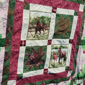 May include: A patchwork quilt with a horse theme. The quilt features squares with images of horses, surrounded by green and burgundy fabric. Some squares have star patterns. The quilt is a mix of textures and colors.