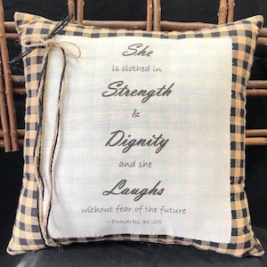 May include: A decorative pillow with a black and tan checkered border. The pillow has a white center with the text "She is clothed in Strength & Dignity and she Laughs without fear of the future -Proverbs 31:25" printed in black.