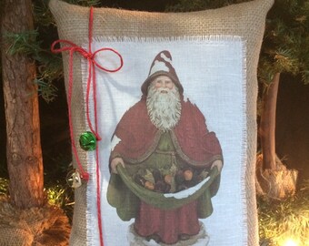 May include: A burlap pillow with a red ribbon and bells. The pillow has a white fabric insert with a printed image of a Santa Claus holding a sack of gifts.
