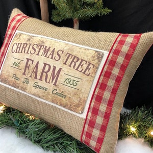 May include: A burlap pillow with red and white checkered trim features a vintage-style sign that reads "Christmas Tree Farm est. 1935 Pine Fir Spruce Cedar".