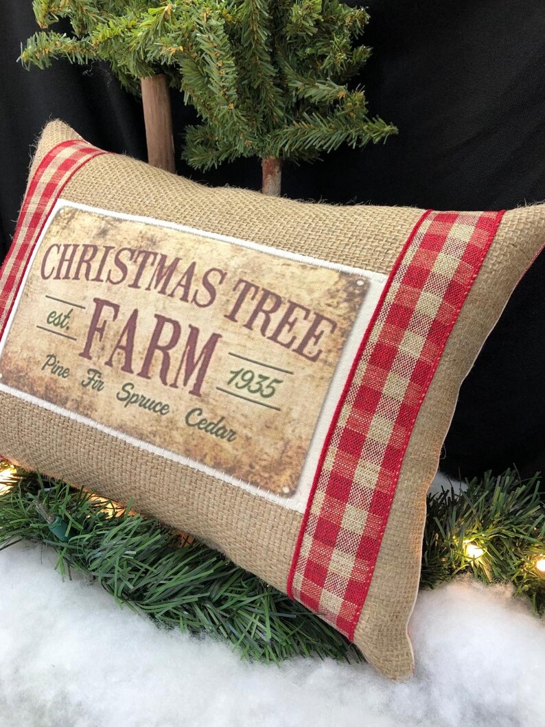 May include: A burlap pillow with red and white checkered trim features a vintage-style sign that reads "Christmas Tree Farm est. 1935 Pine Fir Spruce Cedar".