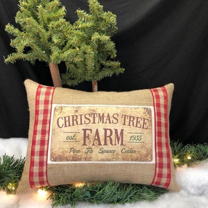 May include: A burlap pillow with red and white gingham trim features a vintage-style sign that reads "Christmas Tree Farm est. 1935 Pine Fir Spruce Cedar".