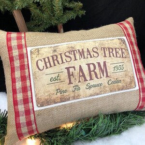 May include: A burlap pillow with red and white checkered trim features a vintage-style sign that reads "Christmas Tree Farm est. 1935 Pine Fir Spruce Cedar".