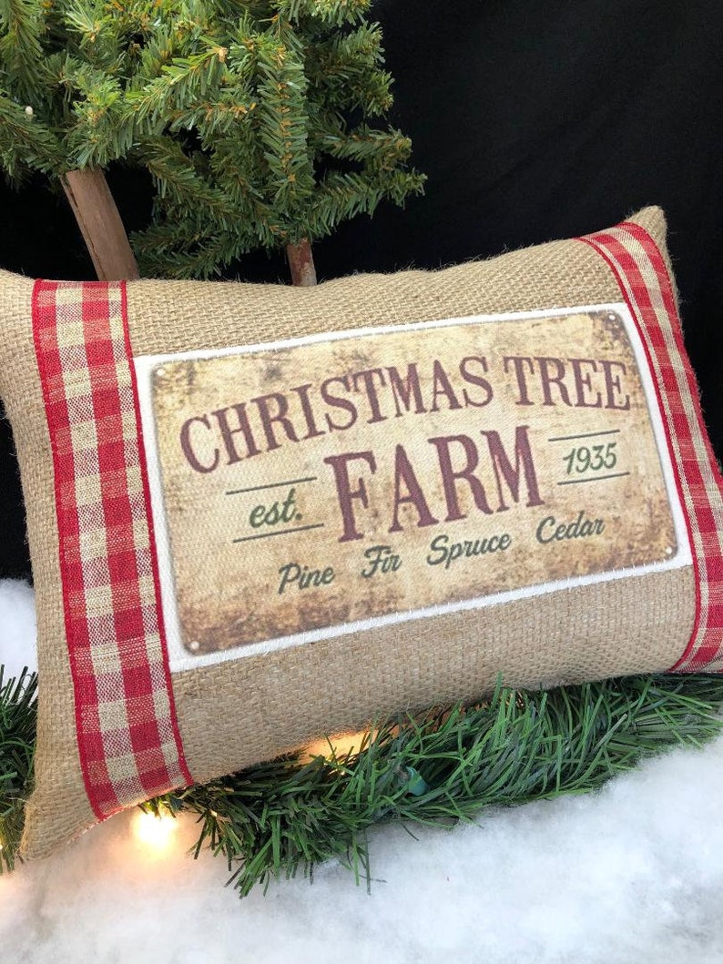 May include: A burlap pillow with red and white checkered trim features a vintage-style sign that reads "Christmas Tree Farm est. 1935 Pine Fir Spruce Cedar".