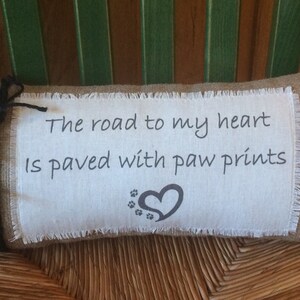 May include: A burlap pillow with a white fabric insert featuring the text "The road to my heart is paved with paw prints" and a black heart with paw prints.
