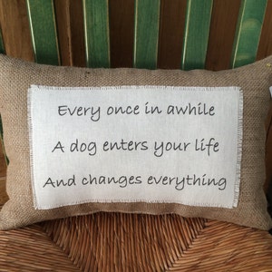 May include: A burlap pillow with white fabric insert featuring the text "Every once in awhile A dog enters your life And changes everything".
