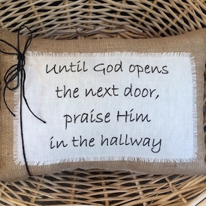 May include: A burlap pillow with a white fabric insert featuring the text "Until God opens the next door, praise Him in the hallway".