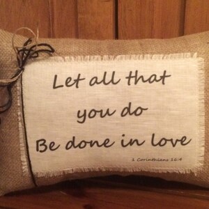 Pode incluir: Uma almofada de juta com um inserto de tecido branco com o texto "Let all that you do Be done in love 1 Corinthians 16:14".