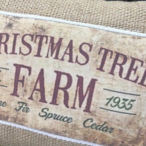 May include: A burlap Christmas tree farm sign with a vintage look. The sign is brown with white stitching around the edges. The text on the sign reads "Christmas Tree Farm 1935" with the words "Pine Fir Spruce Cedar" underneath.