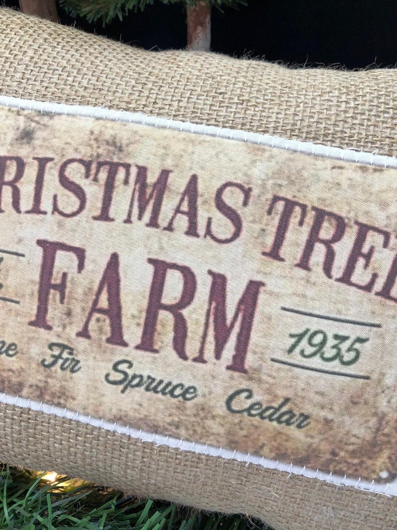 May include: A burlap Christmas tree farm sign with a vintage look. The sign is brown with white stitching around the edges. The text on the sign reads "Christmas Tree Farm 1935" with the words "Pine Fir Spruce Cedar" underneath.