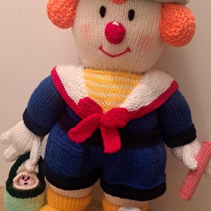 May include: A knitted toy clown with a green hat, red bow, and blue and white sailor shirt. The clown is holding a green bucket and a pink stick. The clown is wearing yellow boots with a pink crab on one foot.