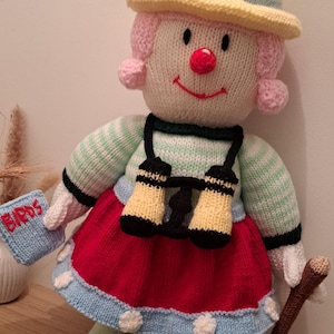 May include: A knitted doll wearing a yellow hat with a green and white pattern, a red skirt, and a green and white striped shirt. The doll is holding binoculars and a stick. The doll is standing on a wooden surface.