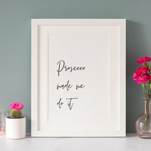Op de afbeelding: Witte ingelijste print met zwarte kalligrafie die "Prosecco made me do it" luidt. De print staat op een wit oppervlak met een roze cactus in een witte pot en een roze glazen vaas met roze bloemen.