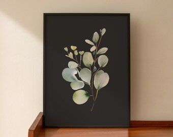 May include: A framed botanical print featuring a watercolor illustration of eucalyptus leaves and branches. The artwork showcases soft green and blue-green foliage against a dark background. The frame is black, and the print is displayed on a wooden surface.
