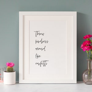 Könnte beinhalten: Weißer gerahmter Druck mit schwarzem Kalligraphietext, der "Throw kindness around like confetti" lautet. Der Druck befindet sich auf einer weißen Oberfläche mit einem rosa Kaktus in einem weißen Topf und einer rosa Glasvase mit rosa Blumen.
