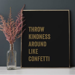May include: A black framed sign with gold lettering that reads "Throw Kindness Around Like Confetti". The sign is sitting on a white surface in front of a grey wall. A vase with pink flowers is to the left of the sign.