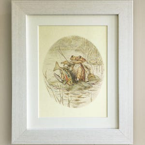 May include: A framed illustration of two frogs sitting on a lily pad in a pond. The frogs are wearing clothes and holding fishing rods. One frog has caught a fish.
