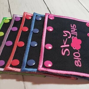 Op de afbeelding: Een stapel geborduurde zwarte stoffen patches met kleurrijke randen en drukknoopsluitingen. Elke patch heeft een andere kleur rand, waaronder groen, paars, rood, blauw en roze. De roze omrande patch heeft de naam "Sky" en een telefoonnummer geborduurd.