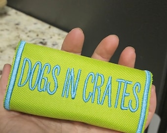 May include: A rectangular dog toy with the text "DOGS IN CRATES" embroidered in light blue on a bright green background. The edges are trimmed with light blue stitching.
