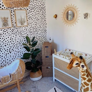 K&ouml;nnte beinhalten: Ein Kinderzimmer mit einer wei&szlig;en Tapete mit schwarzen Punkten, einer Wiege aus Weidengeflecht, einer Wickelkommode mit einer wei&szlig;en Schublade aus braunem Stoff und einem Stofftier in Form einer Giraffe.