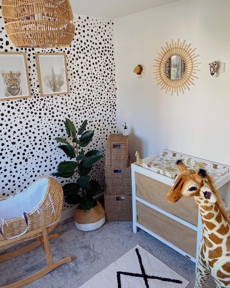 K&ouml;nnte beinhalten: Ein Kinderzimmer mit einer wei&szlig;en Tapete mit schwarzen Punkten, einer Wiege aus Weidengeflecht, einer Wickelkommode mit einer wei&szlig;en Schublade aus braunem Stoff und einem Stofftier in Form einer Giraffe.
