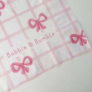 May include: White fabric featuring a pink grid pattern and pink watercolour bows. The text "Bobbin & Bumble" is printed in pink. The fabric has a slightly creased appearance.