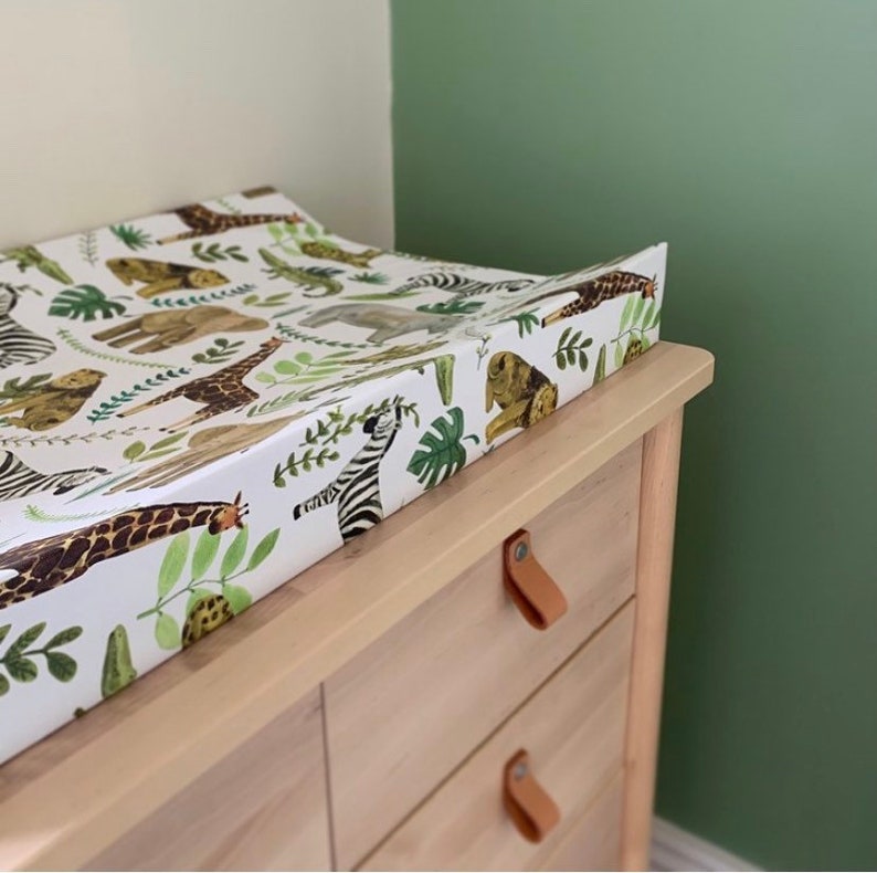 K&ouml;nnte beinhalten: Eine wei&szlig;e Wickelauflage mit einem Dschungeltier-Druck mit Giraffen, Zebras, Elefanten und L&ouml;wen. Die Wickelauflage liegt auf einer Wickelkommode aus hellem Holz mit Leder-Schubladengriffen.