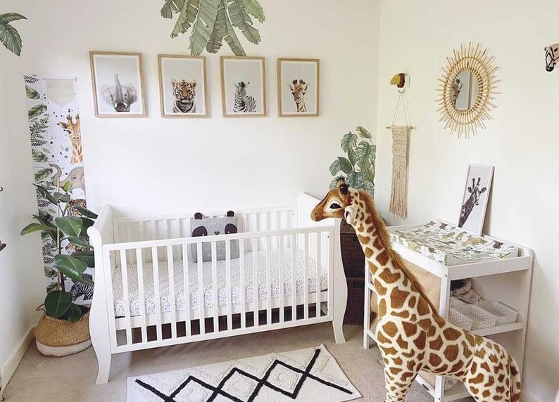 K&ouml;nnte beinhalten: Ein wei&szlig;es Babybett mit einer wei&szlig;en und schwarzen gemusterten Matratze, einem Stofftier-Giraffe und einer Wickelkommode mit einer wei&szlig;en und schwarzen gemusterten Wickelauflage. Das Zimmer ist im Dschungel-Stil eingerichtet, mit gr&uuml;ner und brauner Tiermuster-Tapete und gerahmten Tierdrucken an der Wand.