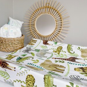 K&ouml;nnte beinhalten: Eine wei&szlig;e Wickelauflage mit einem Dschungeltier-Druck, der eine Giraffe, einen Elefanten, ein Zebra, ein Nashorn, einen Leoparden, einen Alligator und Palmbl&auml;tter zeigt. Die Wickelauflage liegt auf einer wei&szlig;en Oberfl&auml;che mit einem Weidenkorb und einem Sonnenstrahlenspiegel im Hintergrund.