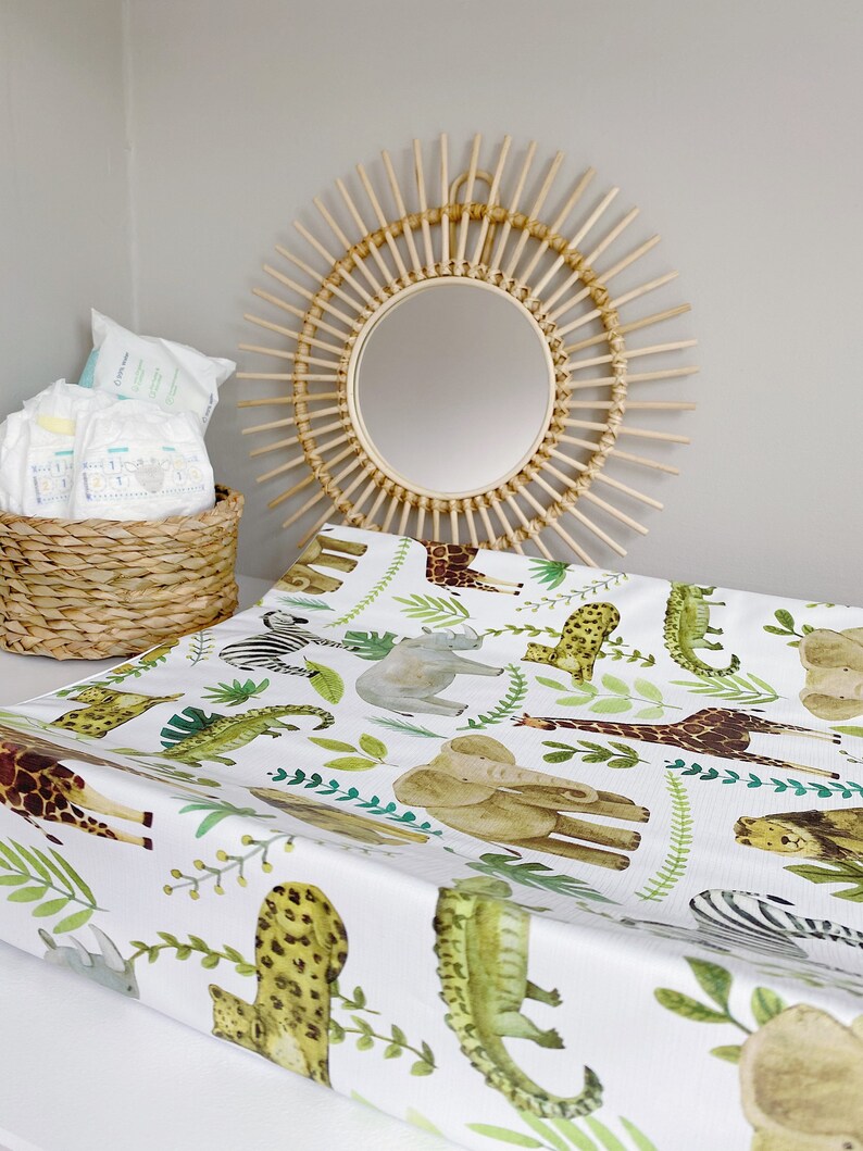 K&ouml;nnte beinhalten: Eine wei&szlig;e Wickelauflage mit einem Dschungeltier-Druck, der eine Giraffe, einen Elefanten, ein Zebra, ein Nashorn, einen Leoparden, einen Alligator und Palmbl&auml;tter zeigt. Die Wickelauflage liegt auf einer wei&szlig;en Oberfl&auml;che mit einem Weidenkorb und einem Sonnenstrahlenspiegel im Hintergrund.