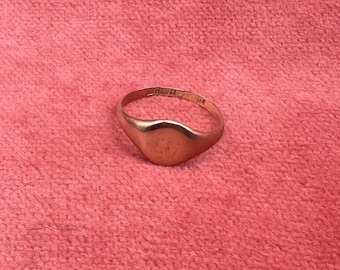 May include: A gold signet ring with a smooth, rounded face. The ring is set against a pink background. The band of the ring has some text. The ring is a classic piece of jewelry.