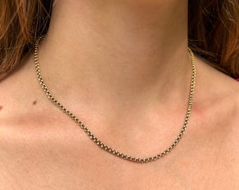 May include: A gold chain necklace featuring a series of small, uniform, rounded interlocking links. The necklace rests against the neck, displaying a polished metallic finish and a consistent, circular link pattern throughout the entire length of the chain.