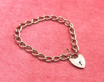 May include: A gold-colored chain bracelet with a heart-shaped padlock charm. The bracelet features a series of oval links and a small chain connecting the padlock. The padlock has a keyhole design. The bracelet is displayed on a pink surface.